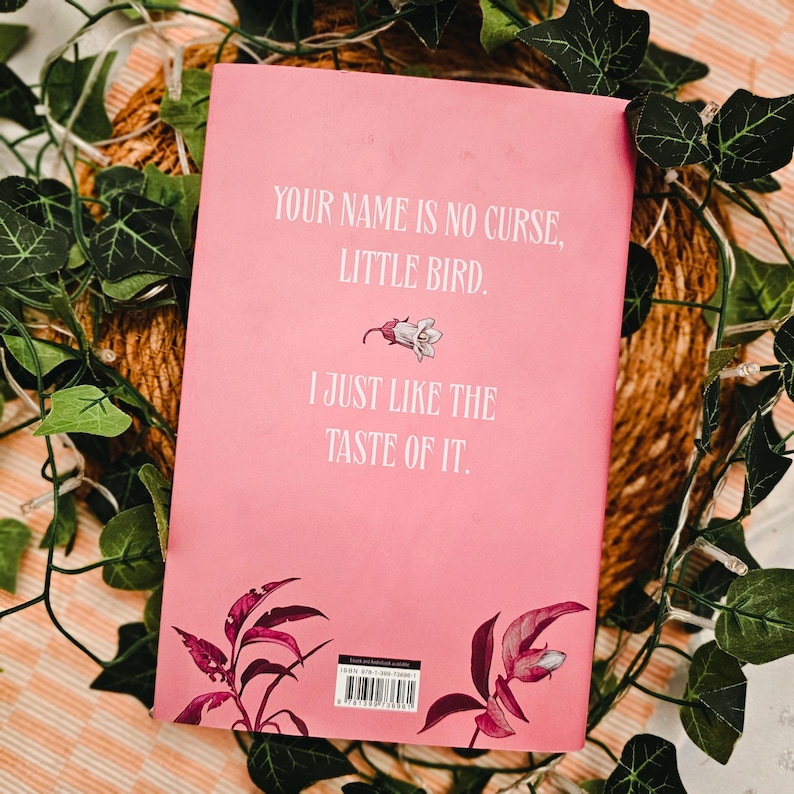 May include: A pink book cover with the text "YOUR NAME IS NO CURSE, LITTLE BIRD. I JUST LIKE THE TASTE OF IT." The cover features a floral illustration and is surrounded by green leaves and a woven background.