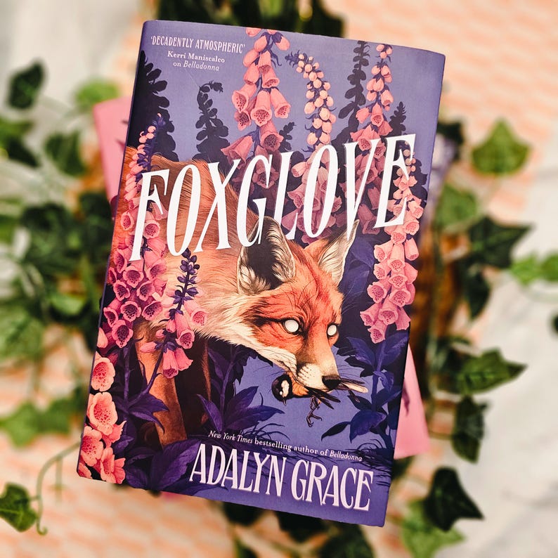 May include: A hardcover book titled "FOXGLOVE" by Adalyn Grace. The cover features a red fox with white eyes amidst purple and pink flowers. The text "DECADENTLY ATMOSPHERIC" and "New York Times bestselling author of Belladonna" are also visible.