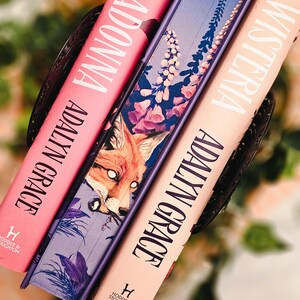 May include: Three hardcover books with titles "Belladonna", "Wisteria", and a third book with a fox illustration. The books are in shades of pink, purple, and peach. The author's name "Adalyn Grace" is printed on the spines.