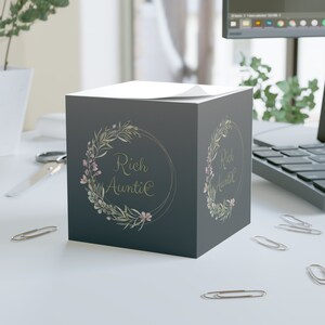 Op de afbeelding: Een grijze, kubusvormige notitieblok met een bloemenkransontwerp en de tekst "Rich Auntie" in gouden letters.
