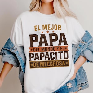 May include: White t-shirt with the text "EL MEJOR PAPA DEL MUNDO Y EL PAPACITO DE MI ESPOSA" in brown and gold lettering. The shirt has a crew neck and short sleeves. The text is arranged in a stacked format with star accents.