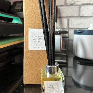 May include: A clear glass bottle with a silver lid and black reeds. The bottle contains a yellow liquid and has a label that reads "Made with love & personalised craft. Vanilla reed diffuser."