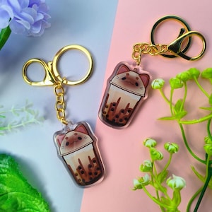 Può includere: Due portachiavi con un design a cartoni animati di tè boba con una faccia di gatto. I portachiavi hanno hardware color oro e sono ambientati su uno sfondo rosa e azzurro. Il tè boba è marrone e beige.