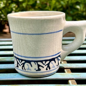 May include: Cream-colored ceramic mug with a crackle glaze and a unique handle design. The mug features two blue horizontal stripes and a decorative blue pattern near the base. A handmade, rustic style mug.