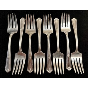 May include: A collection of eight silver forks with a classic design. Each fork features a tined head and a decorative handle with ornate detailing. The forks are arranged on a black surface, showcasing their polished finish.