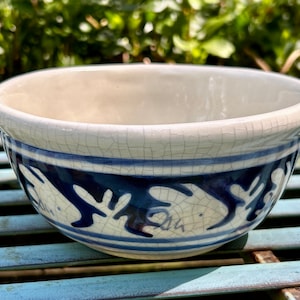 May include: A ceramic bowl with a cream-colored interior and a blue and white decorative band. The band features a stylized design in dark blue. The bowl has a crackled glaze finish, adding to its vintage appeal.