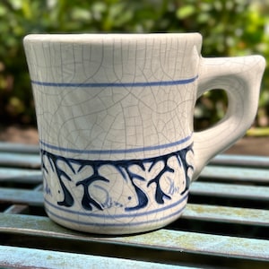 May include: A white ceramic mug with a crackle glaze finish. The mug features blue horizontal stripes and a decorative blue pattern around the base. The handle is a unique, curved design.
