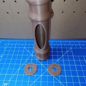 May include: A brown, 3D-printed object with a cylindrical body and an oval cutout. Two brown circular washers are also present. The object appears to be a component or part of a larger assembly. The background is a blue cutting mat.