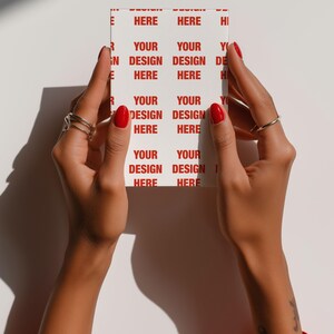 May include: A white card with the words "YOUR DESIGN HERE" repeated in red. The card is held by hands with red nail polish and silver rings. The person is wearing a red top.