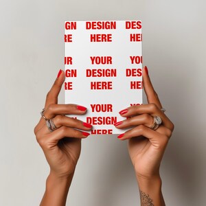May include: A white rectangular card with red text that reads "DESIGN HERE" and "YOUR DESIGN HERE". The card is held up by hands with red nail polish and multiple rings.