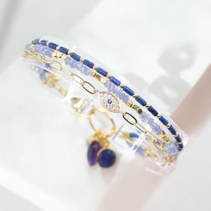 Può includere: Un braccialetto a catena dorata con tre fili di perline blu e dorate. Il braccialetto presenta un ciondolo con un occhio blu malocchio e due pietre blu e viola pendenti.