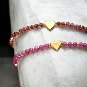 May include: Two delicate bracelets with pink gemstone beads and gold heart charms. The bracelets are stacked on a white surface.
