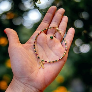 Könnte beinhalten: Ein goldfarbenes Perlenarmband mit einem grünen Edelstein-Anhänger und einem goldenen Buchstaben 'N'-Anhänger. Das Armband besteht aus kleinen, bunten Perlen.