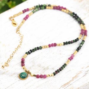 May include: A necklace with a gold chain and a green stone pendant. The necklace is decorated with small beads in pink, green, and black, and gold-coloured square beads. The necklace has a gold clasp. The necklace is approximately 40 cm in length.