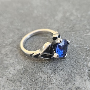May include: A silver ring with a large, round, deep blue gemstone. The ring has an intricate design with decorative elements on the band and prongs holding the stone. The ring is set against a gray background.