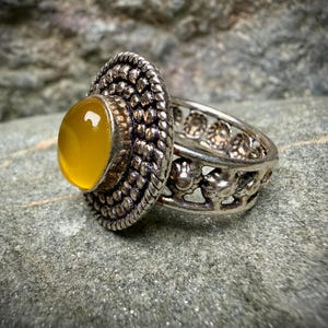 May include: A silver ring with a large, oval, yellow gemstone. The ring has a detailed, textured band and a decorative bezel around the stone. The ring is set against a textured, gray background.