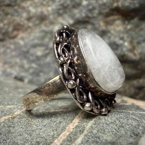 A silver ring with a large white stone, possibly a gemstone, is displayed against a blurred background.