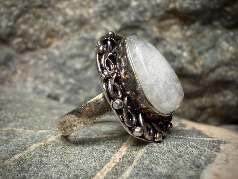 A silver ring with a large white stone, possibly a gemstone, is displayed against a blurred background.