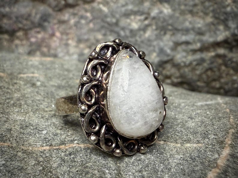 A silver ring with a large, white gemstone, possibly a moonstone, set in an ornate, decorative metal frame.