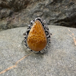 May include: A silver ring featuring a teardrop-shaped, amber-colored stone with a textured surface. The stone is set in a decorative silver band with a detailed border of small circles and curved accents. The ring is displayed on a gray stone surface.