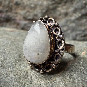 A silver ring with a large, white stone or gemstone in the center, surrounded by an intricate, decorative metal frame.