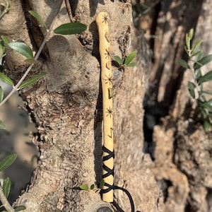 Puede incluir: Una varita de madera clara con diseños de estrellas y líneas talladas, acentuada con pequeñas piedras. La parte inferior está envuelta con un cordón negro. La varita mide aproximadamente 30 cm de largo.