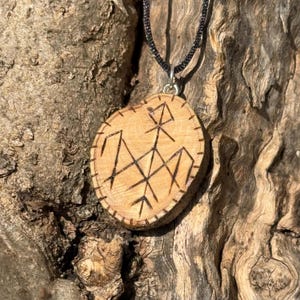 May include: A wooden pendant necklace with a black cord. The pendant is a round piece of wood with a carved symbol in the center. The symbol is a geometric design with lines and angles.