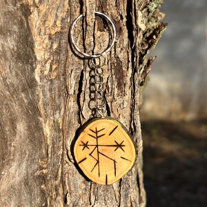 Puede incluir: Un llavero de madera con un símbolo tallado, unido a un anillo y una cadena plateados. El llavero está colgado de un tronco de árbol.