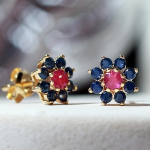 Può includere: Un paio di orecchini a lobo in oro a forma di fiore. Ogni orecchino presenta una pietra preziosa rossa rotonda centrale circondata da otto piccole pietre blu scuro rotonde disposte in un motivo circolare, creando un vivace design floreale.