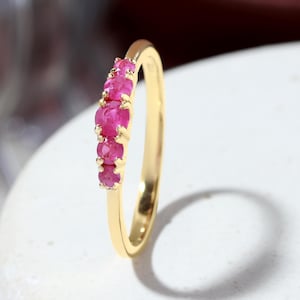 Op de afbeelding: Een dunne ring van gepolijst geelgoud met een rij van vijf kleine, ronde, felroze robijnen. De stenen zijn in een verticale lijn geplaatst en worden op hun plek gehouden door kleine metalen pootjes, wat zorgt voor een verfijnde uitstraling.
