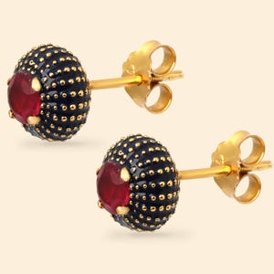 May include: A pair of stud earrings featuring a round, dark blue, textured dome with a central, faceted, circular red gemstone. The earrings have a gold-toned post and a matching gold-toned butterfly clutch backing, providing a secure fit.