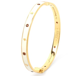 Op de afbeelding: Een goudkleurige scharnierende armband met een witte emaille inleg op de buitenkant, versierd met kleine gouden schoppensymbolen en ingelegde ronde bruine stenen. De binnenkant is gegraveerd met de tekst Alice is impossible.