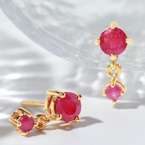 May include: A pair of gold drop earrings featuring two round, vibrant red gemstones on each piece. The design includes a smaller stone at the top and a larger stone suspended below, both set in polished gold metal with a secure post-back closure.