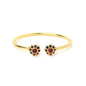 Puede incluir: Una pulsera abierta de tono dorado con dos terminales en forma de flor. Cada terminal tiene una piedra roja redonda en el centro rodeada por un anillo de pequeñas piedras circulares de color azul oscuro, creando un motivo floral.