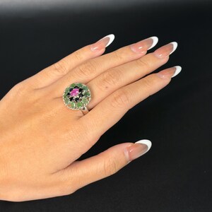 May include: A silver ring with a circular design featuring a central pink gemstone surrounded by black and green gemstones. The ring is set against a black background.