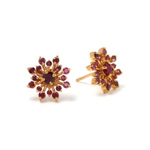 May include: A pair of gold-colored stud earrings. Each earring features a central, round, dark red gemstone surrounded by smaller, lighter red gemstones, creating a starburst design. The earrings are set against a white background.