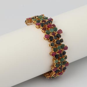 Puede incluir: Una pulsera dorada con un diseño floral. La pulsera está adornada con una variedad de piedras preciosas de colores, incluyendo piedras rojas, verdes y negras. Las piedras preciosas tienen forma de lágrima y están muy juntas.