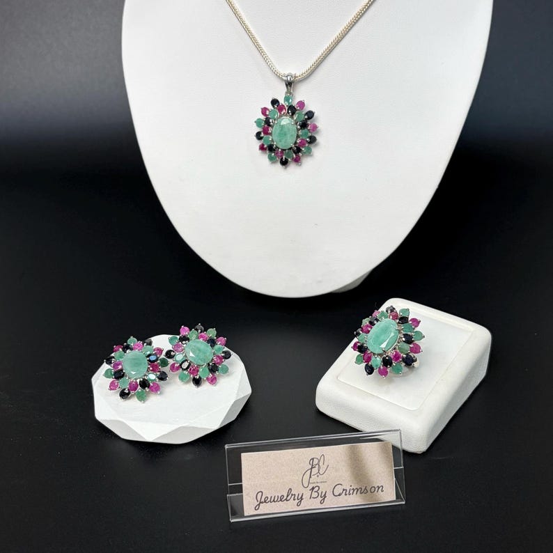 Puede incluir: Un conjunto de joyas que incluye un collar con colgante, pendientes y un anillo. Cada pieza tiene una piedra verde ovalada central rodeada de piedras rojas, negras y verdes m&aacute;s peque&ntilde;as. El conjunto se exhibe sobre pedestales blancos sobre un fondo negro.