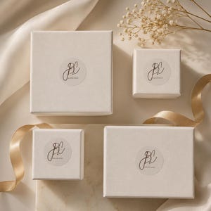 Puede incluir: Cuatro cajas de regalo blancas de diferentes tama&ntilde;os est&aacute;n dispuestas sobre una superficie color crema. Cada caja tiene una pegatina circular con un logotipo de estilo manuscrito. Una cinta dorada y ramitas de flores secas a&ntilde;aden elementos decorativos.