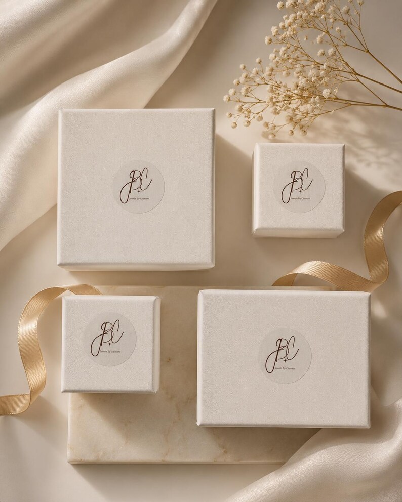 Puede incluir: Cuatro cajas de regalo blancas de diferentes tama&ntilde;os est&aacute;n dispuestas sobre una superficie color crema. Cada caja tiene una pegatina circular con un logotipo de estilo manuscrito. Una cinta dorada y ramitas de flores secas a&ntilde;aden elementos decorativos.