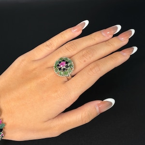 May include: A silver ring featuring a floral design with a central pink gemstone surrounded by green and black stones. The ring is set against a black background, showcasing the vibrant colors of the gemstones. The ring is a unique piece of jewelry.