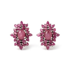 May include: A pair of stud earrings with a central oval ruby, encircled by smaller, teardrop-shaped rubies. The earrings have a silver-coloured metal setting and a floral design. The rubies are a deep pink colour.