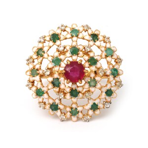 May include: A gold-toned ring featuring a central, round, deep red gemstone surrounded by a floral design. The design incorporates green and white accents, with small, clear gemstones interspersed throughout the floral pattern. The ring is set against a white background.