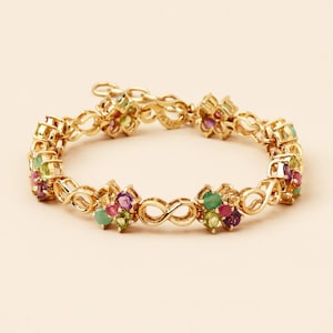 Puede incluir: Pulsera dorada con un diseño floral repetitivo. Cada grupo floral está adornado con pequeñas piedras preciosas verdes, moradas y rojas. La pulsera incorpora símbolos de infinito. Joyería.