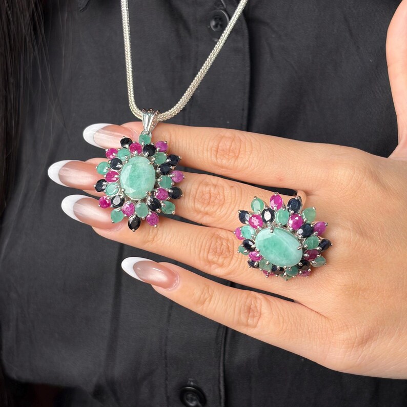 Puede incluir: Un conjunto de collar con colgante y anillo de plata. La joyer&iacute;a presenta una gema grande, ovalada, de color verde claro, rodeada de gemas m&aacute;s peque&ntilde;as en rosa, verde y negro. El collar tiene una cadena de plata.
