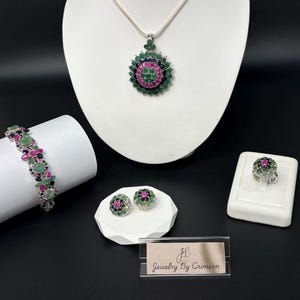 May include: A jewelry set featuring a necklace, bracelet, ring, and earrings. The pieces have a floral design with green, pink, and black gemstones. The set is displayed on white pedestals against a black background. The brand name "Jewelry By Crimson" is visible.