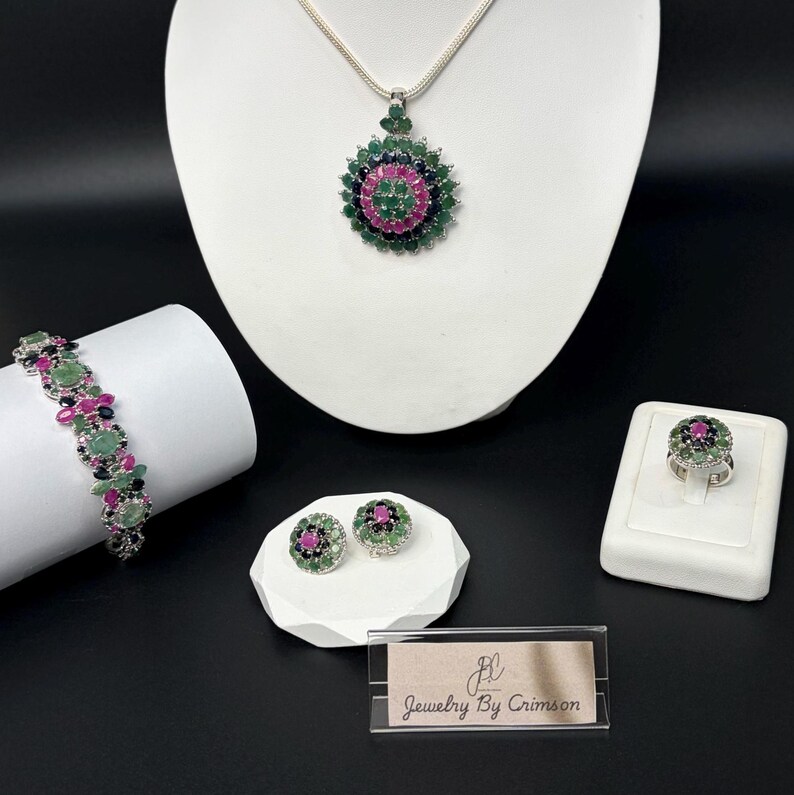 May include: A jewelry set featuring a necklace, bracelet, ring, and earrings. The pieces have a floral design with green, pink, and black gemstones. The set is displayed on white pedestals against a black background. The brand name "Jewelry By Crimson" is visible.