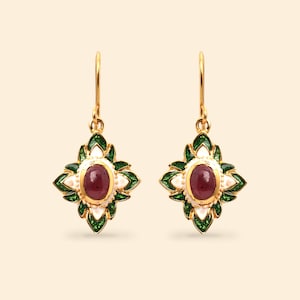 May include: A pair of gold-colored drop earrings. Each earring features a central oval-shaped, dark red gemstone surrounded by a white and green floral design. The earrings have a hook closure.