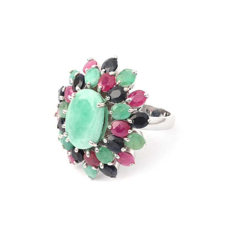 Puede incluir: Un anillo de plata con una gran piedra ovalada de color verde claro en el centro, rodeada de piedras m&aacute;s peque&ntilde;as en forma de l&aacute;grima en tonos verdes, rojos y negros. El anillo tiene una banda pulida y reflectante.