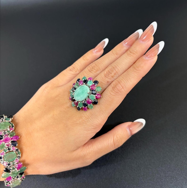 Puede incluir: Un anillo ovalado con una piedra central verde claro, rodeado de piedras m&aacute;s peque&ntilde;as en tonos verdes, rosas y azul oscuro. Tambi&eacute;n se ve una pulsera a juego. La joyer&iacute;a est&aacute; sobre un fondo negro.